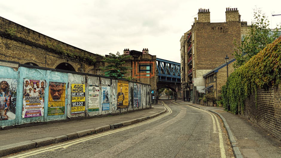 A narrow urban street scene showing a gently curving asphalt road with double yellow lines along the edges, bordered by a low, weathered brick wall on the right side and a taller barrier covered with 