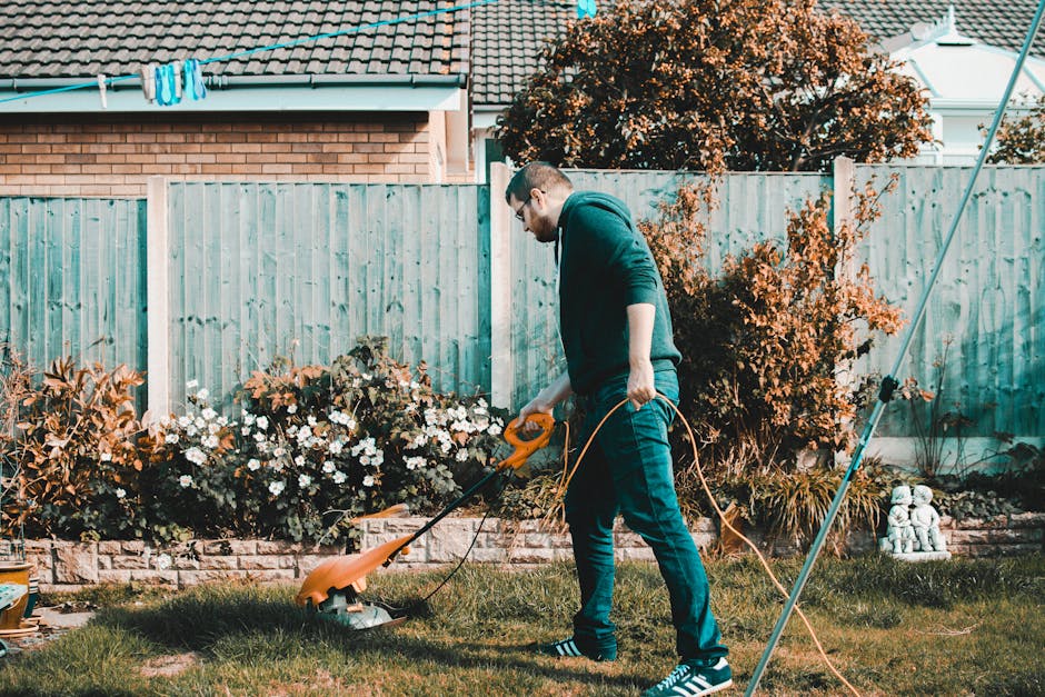 A gardener dressed in a blue waterproof jacket with the hood up, working on a neatly maintained terrace garden. The individual is using a garden rake to clear fallen leaves from a lush, green lawn tha